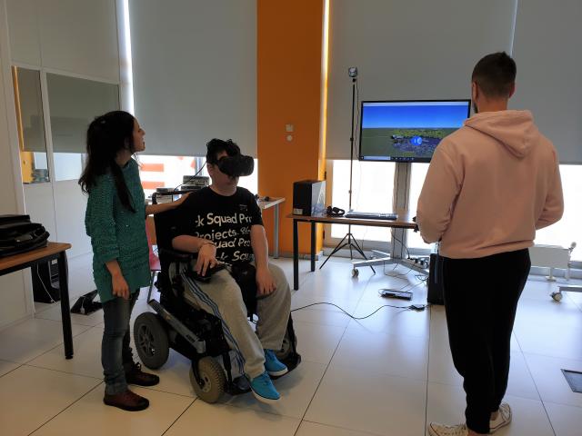 Persona en silla de ruedas usando unos cascos de realidad virtual junto a otras dos personas.